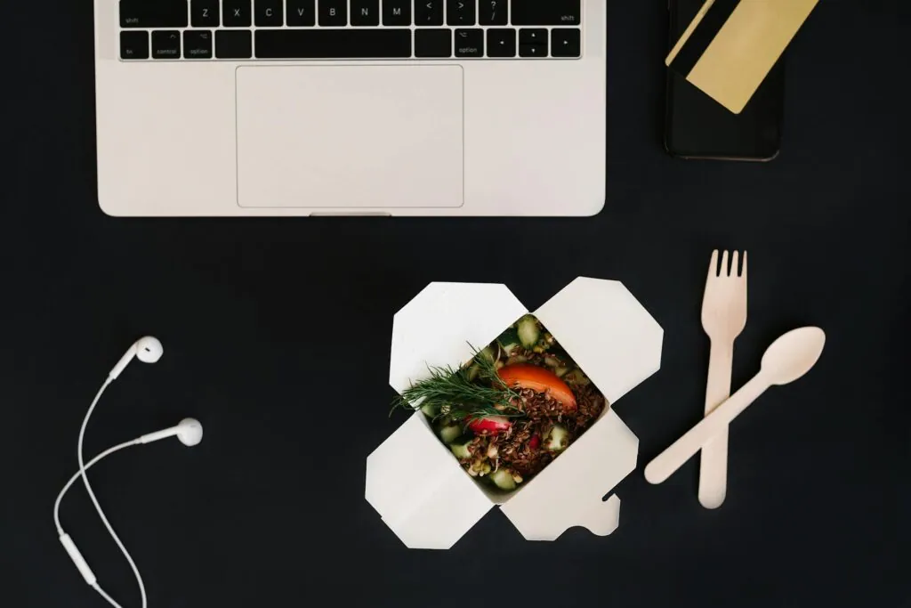 Boîte repas ouverte avec salade et couverts en bois, près d'un ordinateur et d'écouteurs